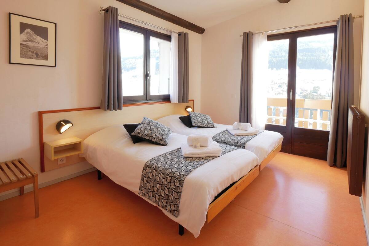 A double bed with patterned pillows, a bedside table with a lamp, and a window with curtains.