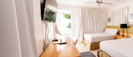 A hotel room with two beds, a desk with a lamp, a ceiling fan, and a window with curtains.
