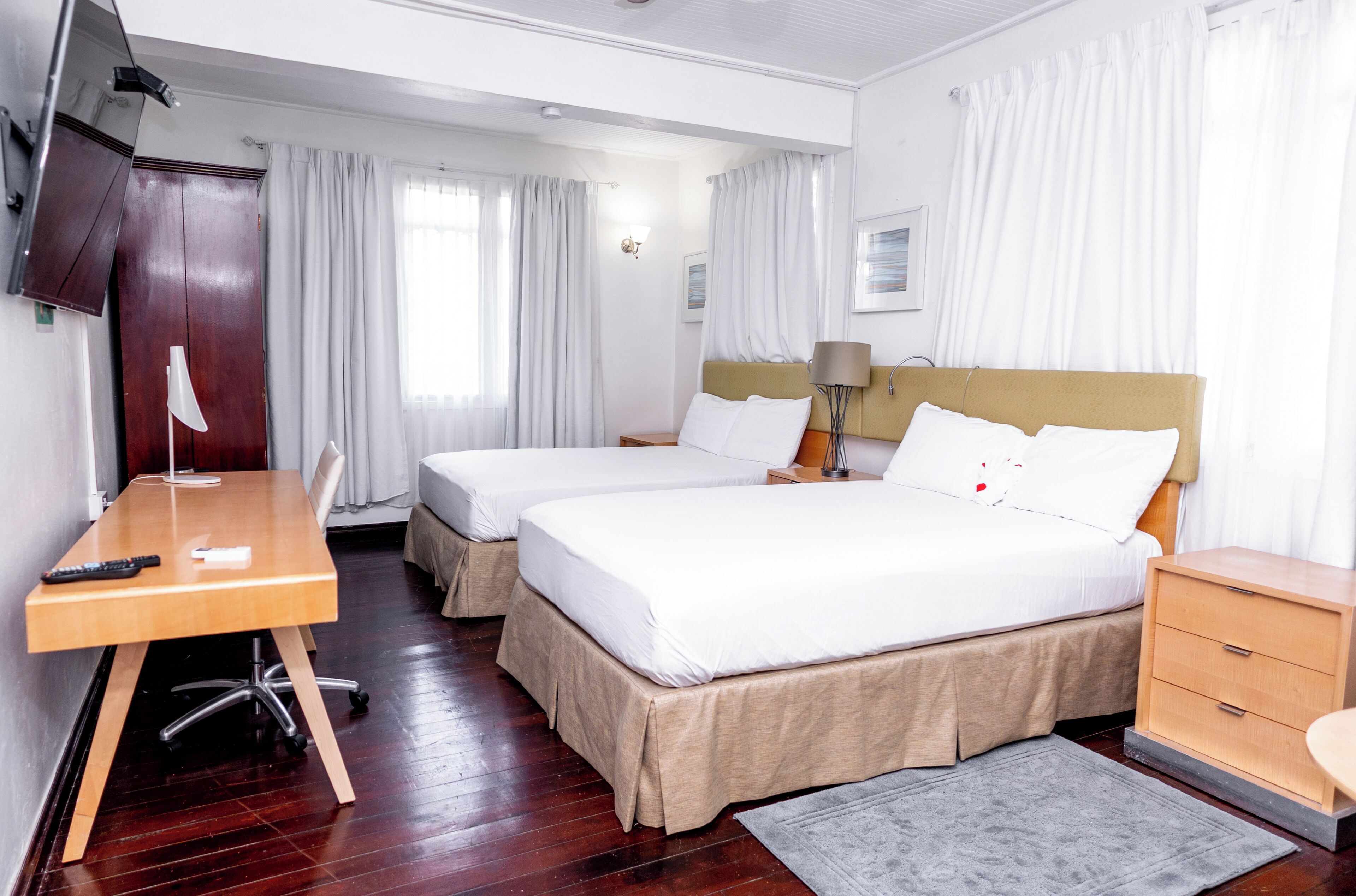 Quarto de hotel com duas camas, uma escrivaninha, televisão e uma pequena mesa de cabeceira com abajur.