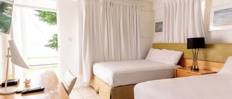 Ein Hotelzimmer mit zwei Betten, einem Schreibtisch mit Lampe, einem Deckenventilator und einem Fenster mit Vorhängen.