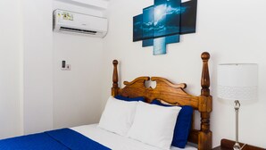 Ein ordentlich bezogenes Bett mit weißer und blauer Bettwäsche, ein hölzerner Kopfteil, eine Wand-Klimaanlage, ein Nachttisch mit Lampe und ein mehrteiliges Kunstwerk darüber.