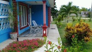 A house with a blue exterior, a porch with a rocking chair, and a garden with various plants and flowers. - Hostal Marisol y Marcos (Ciénaga de Zapata)