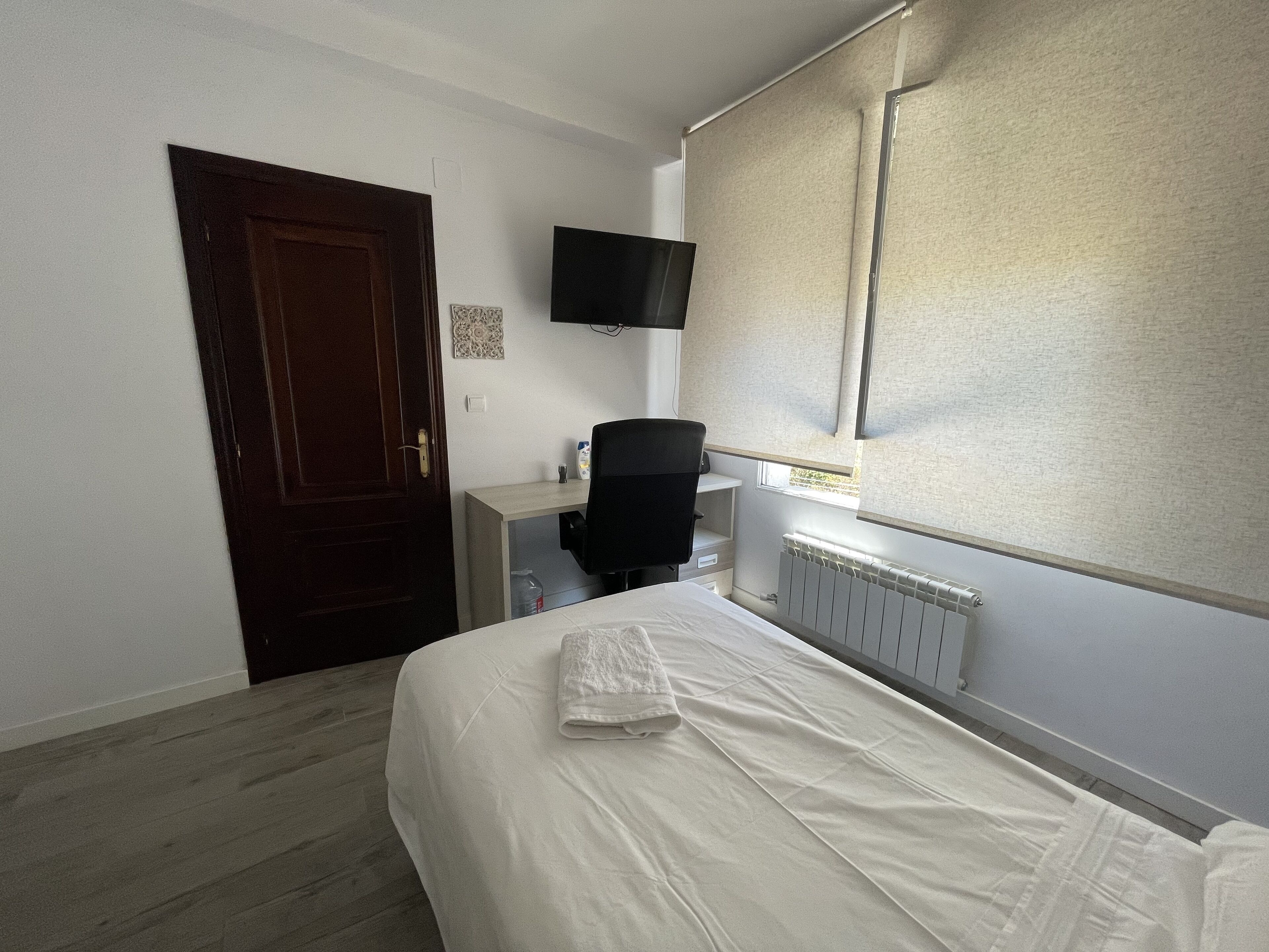 Un dormitorio con cama, escritorio, silla, televisor, puerta y ventana con persianas.