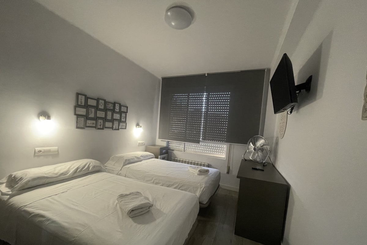 A hotel room with two beds, a flat-screen TV mounted on the wall, and a window with blinds.