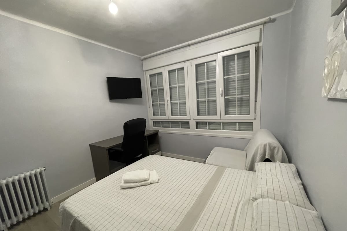 A bedroom with a bed, a desk, a chair, a radiator, and a window with blinds.