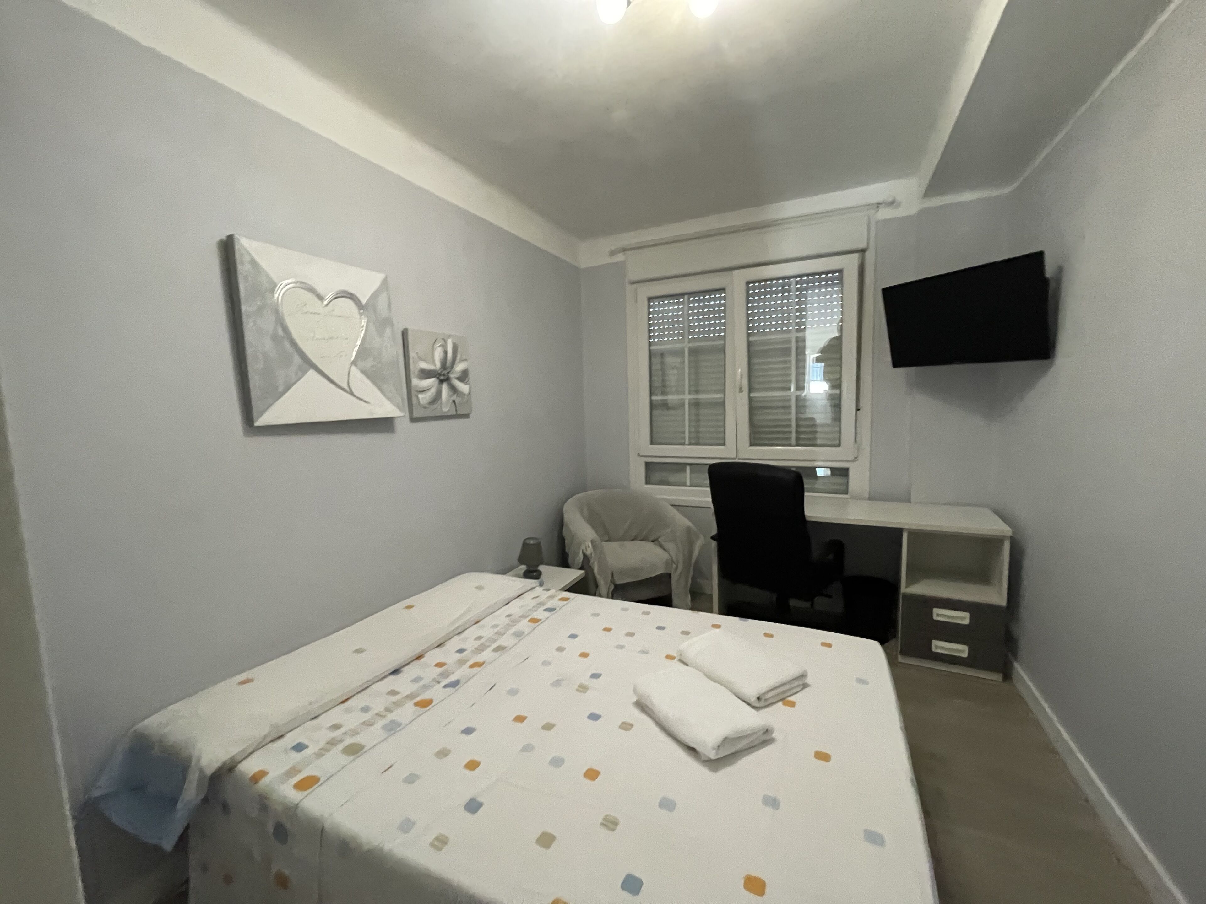 A bedroom with a bed, a desk, a chair, and a TV mounted on the wall.