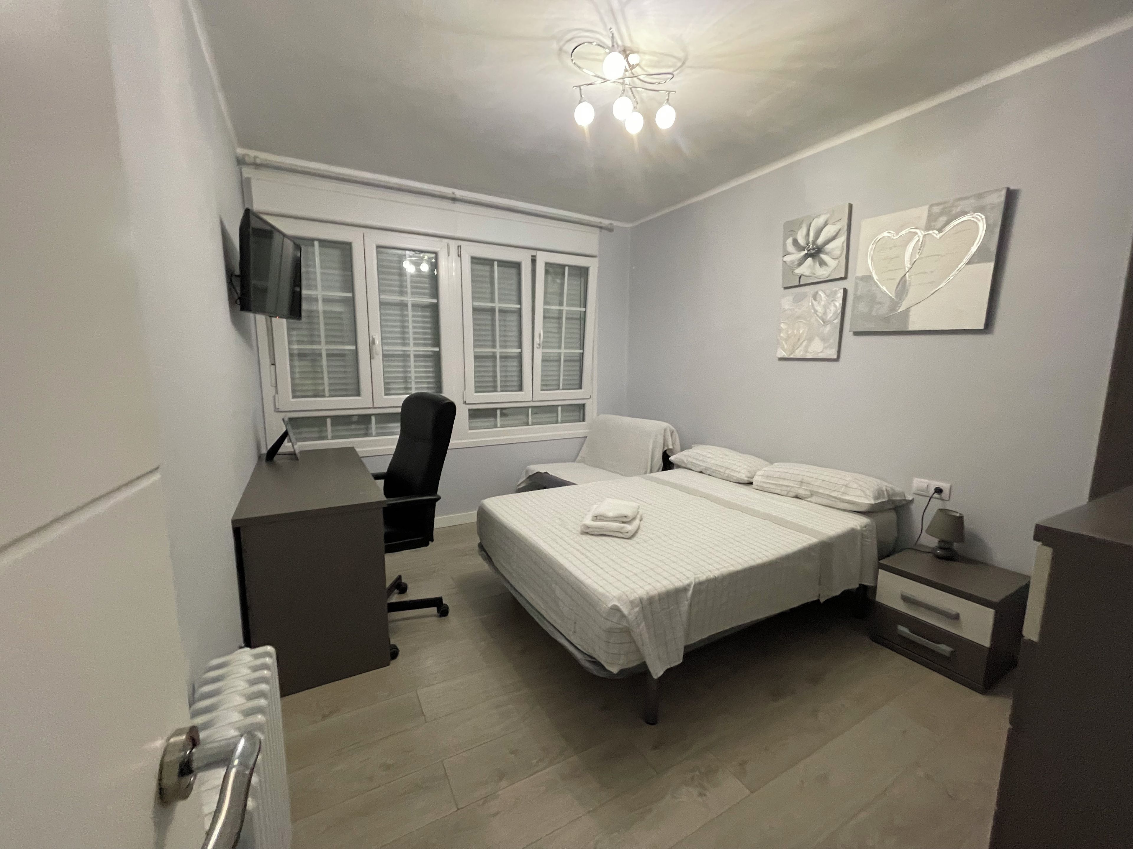 Un dormitorio con cama, escritorio, silla, televisor y obras de arte en la pared.