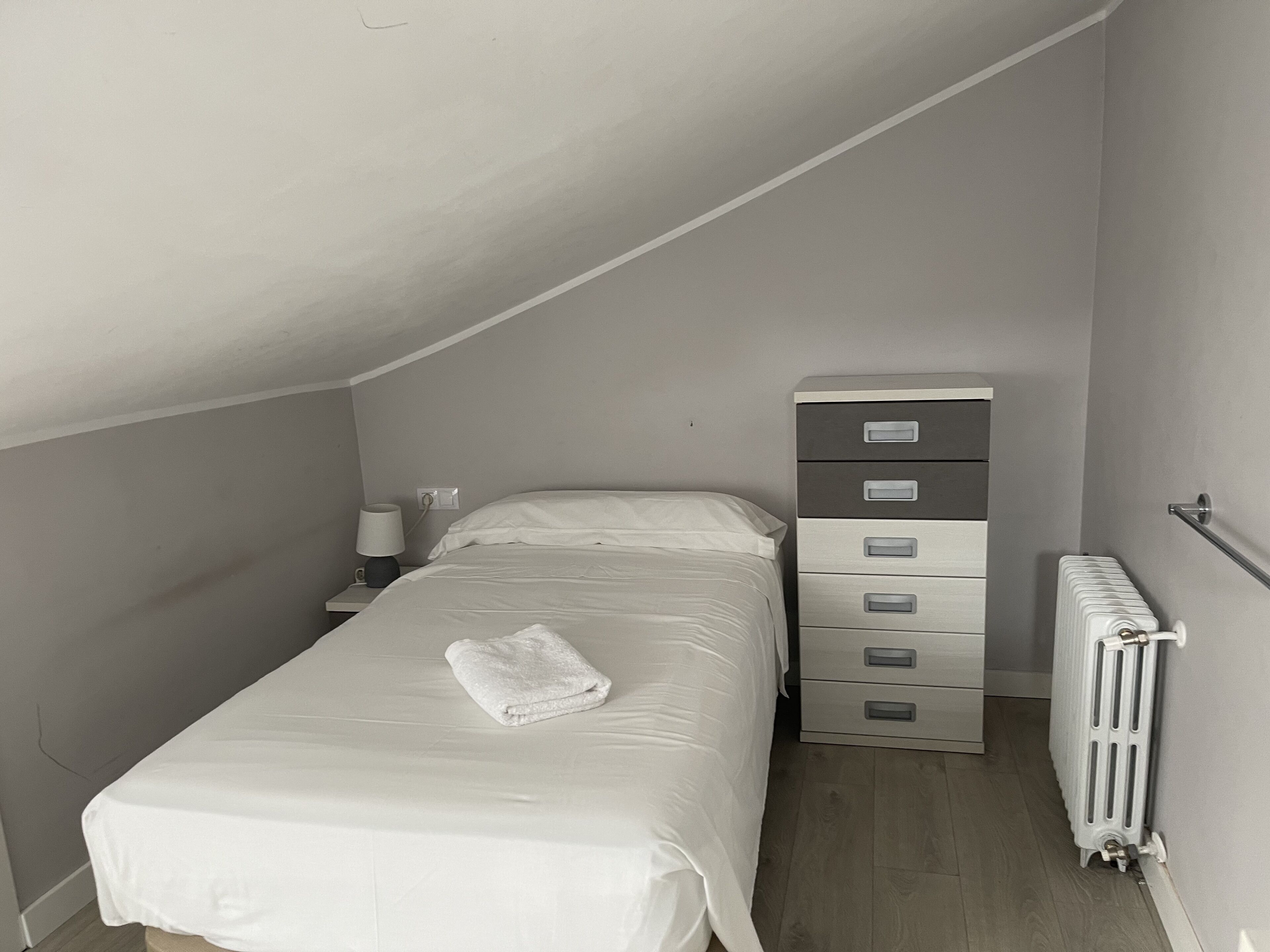 Un dormitorio pequeño y ordenado con una cama individual, una mesita de noche, una lámpara, un aparador y un radiador.