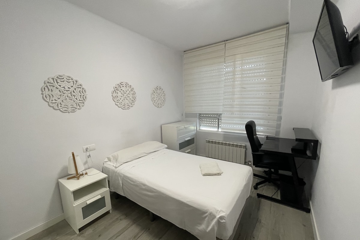 A bedroom with a bed, a desk, a chair, and a TV on the wall.