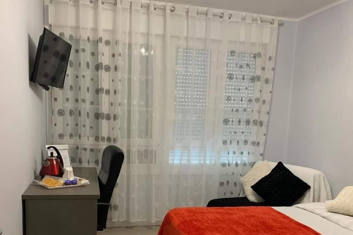 A hotel room with a bed, a desk, a chair, a TV mounted on the wall, and a window with patterned curtains.