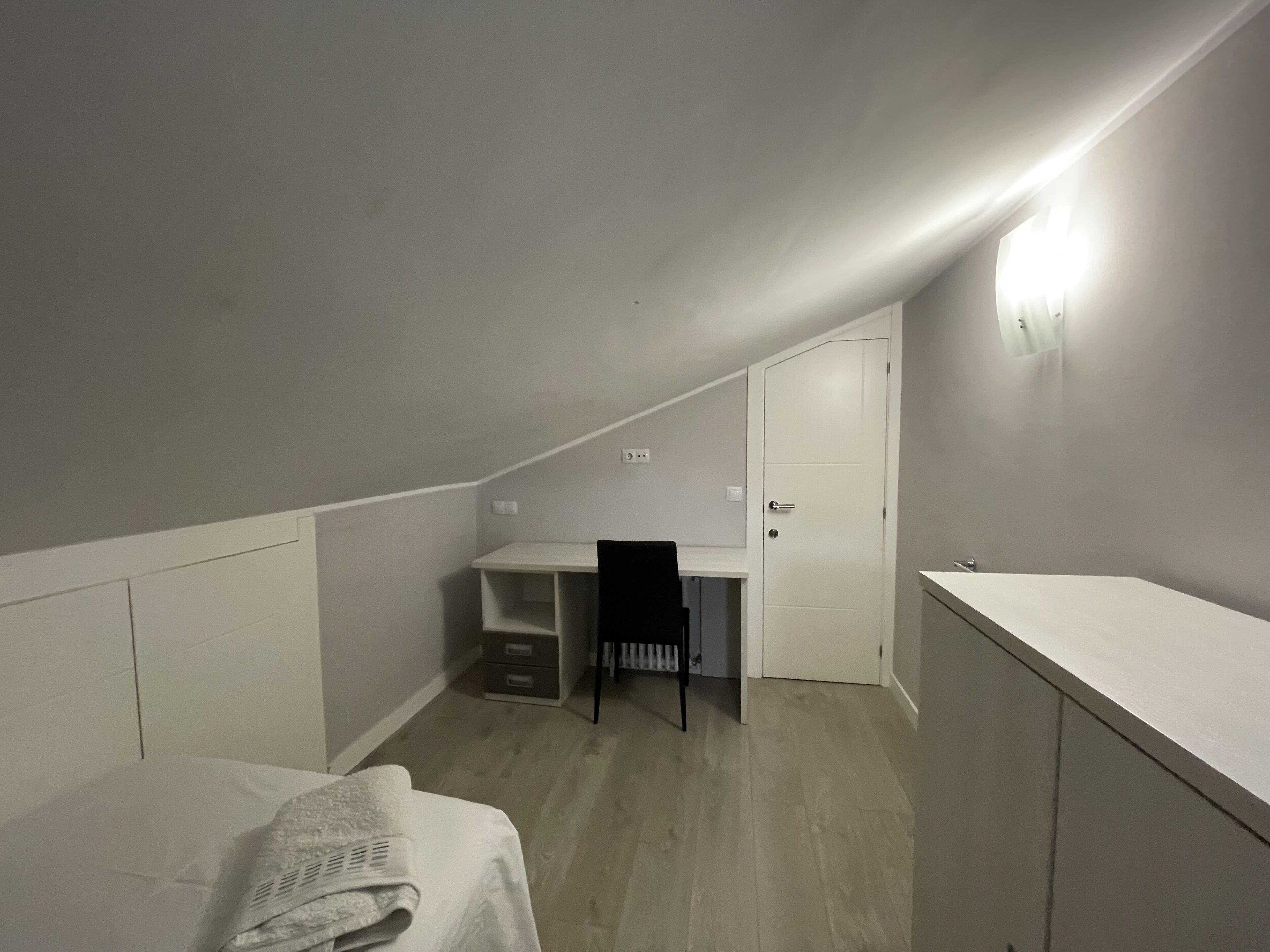 A small, dimly lit attic bedroom with a single bed, a desk, and a chair.