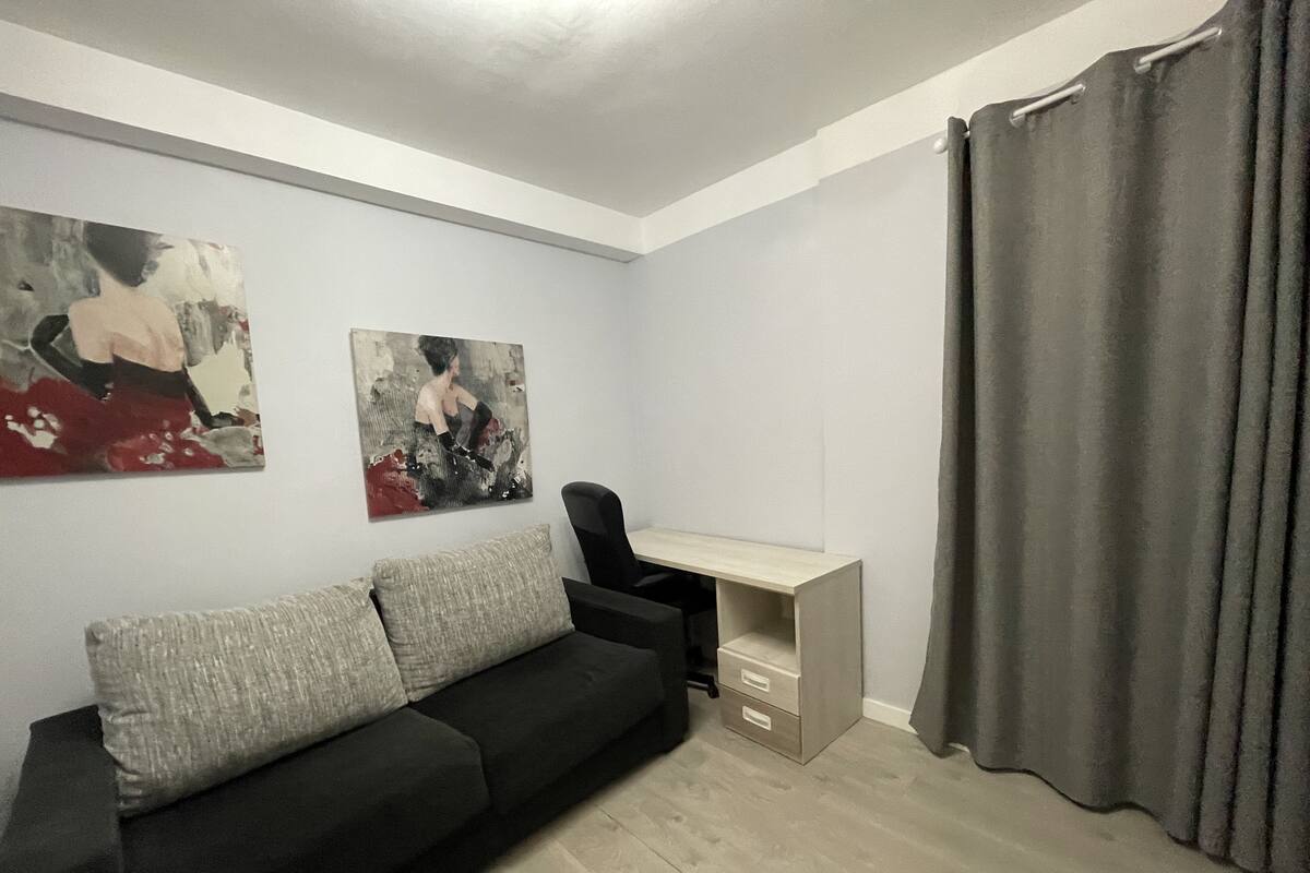 A modern living room with a black sofa, two abstract paintings, a wooden desk, and a grey curtain.
