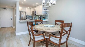 Un comedor con una mesa redonda y cuatro sillas de madera, un candelabro arriba y una cocina al fondo.