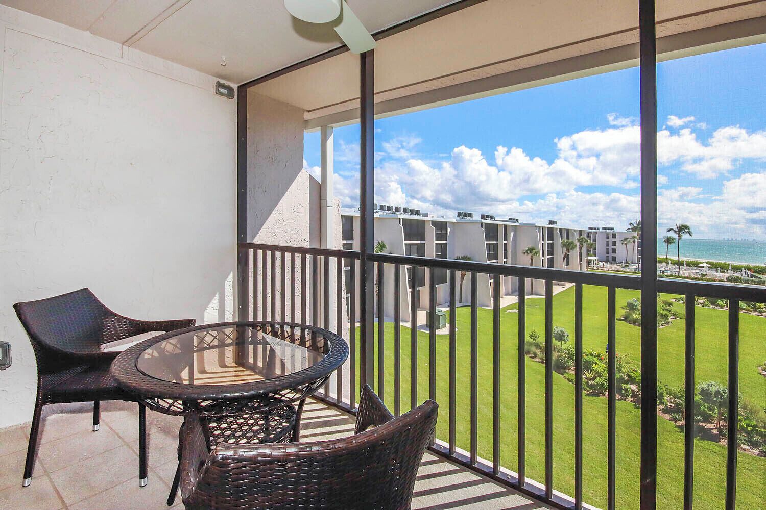 A balcony with a glass table and wicker chairs, overlooking a well-maintained lawn and a building with multiple balconies.