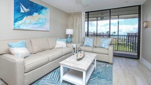 Una sala de estar moderna con un sofá color beige, un sillón a juego y una mesa de centro. Se ve una ventana grande con vistas al exterior, un ventilador de techo y un cuadro de un velero en la pared.