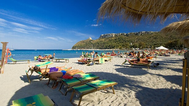 A beach with sun loungers and umbrellas, a clear blue sky, and a hill in the background.