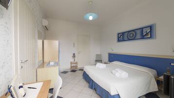 A hotel room with a bed, a desk with a chair, a mirror, a small table with a lamp, and a wall-mounted artwork.