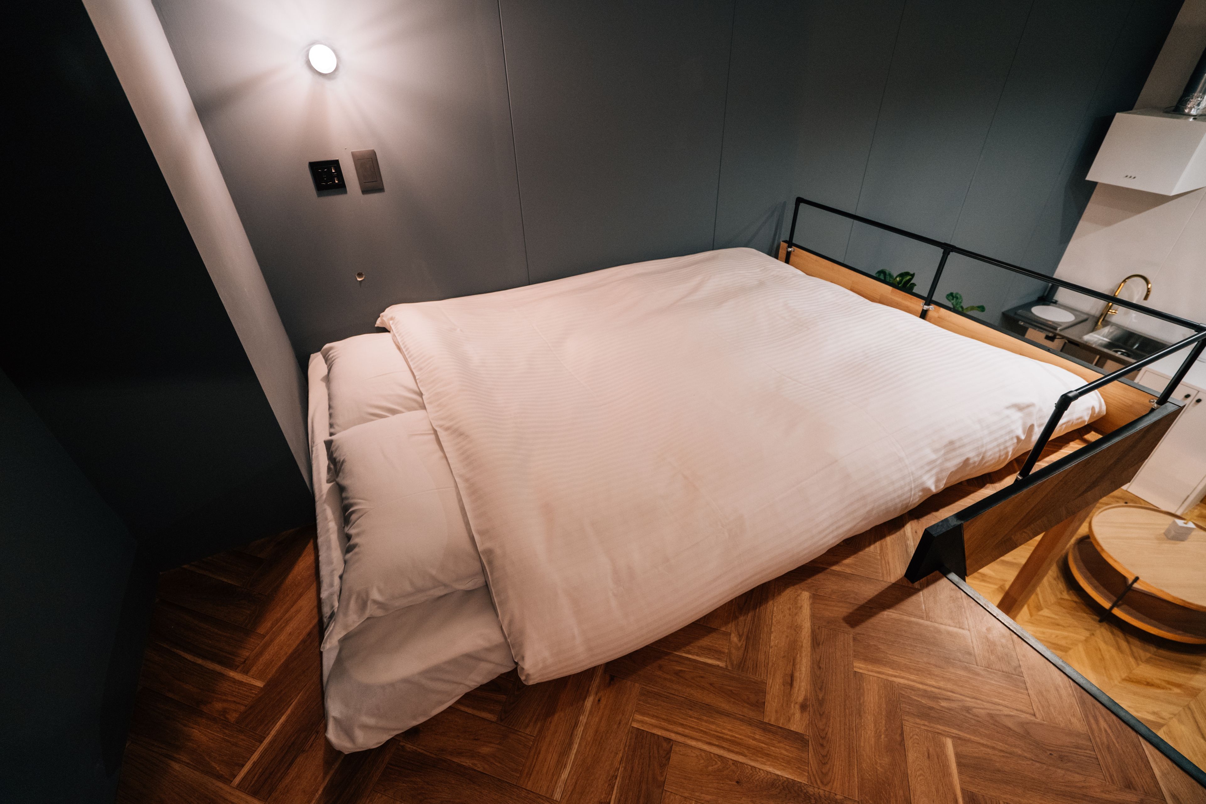 Un lit simple avec un cadre en bois et une tête de lit, une table de chevet avec une lampe, et une petite table ronde avec un tabouret.