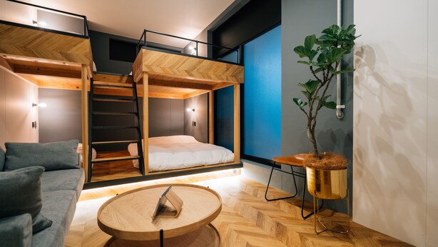 Een moderne kamer met een hoogslaper, een slaapbank, een ronde houten tafel en een potplant.