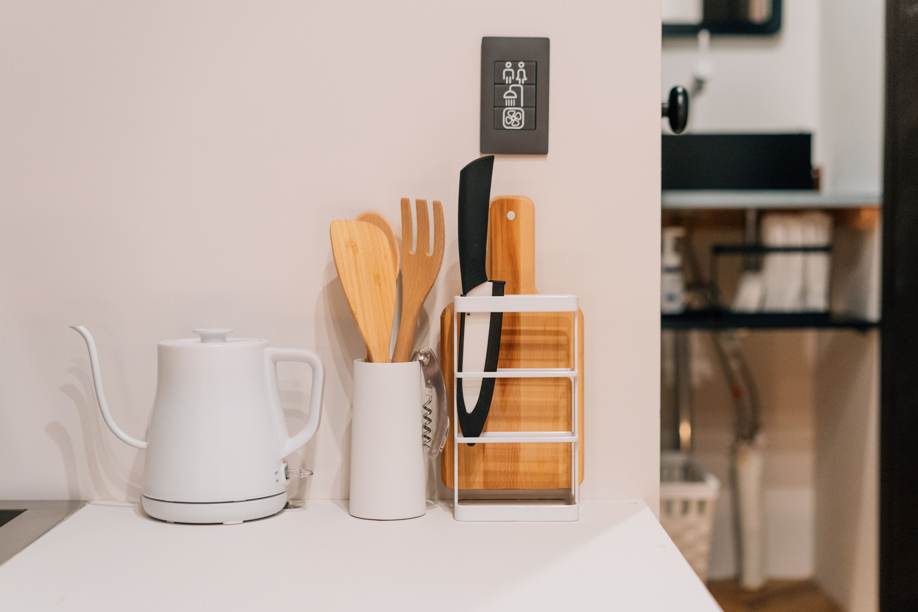 A kitchen counter with a white kettle, a white mug with wooden utensils, and a wooden utensil holder.
