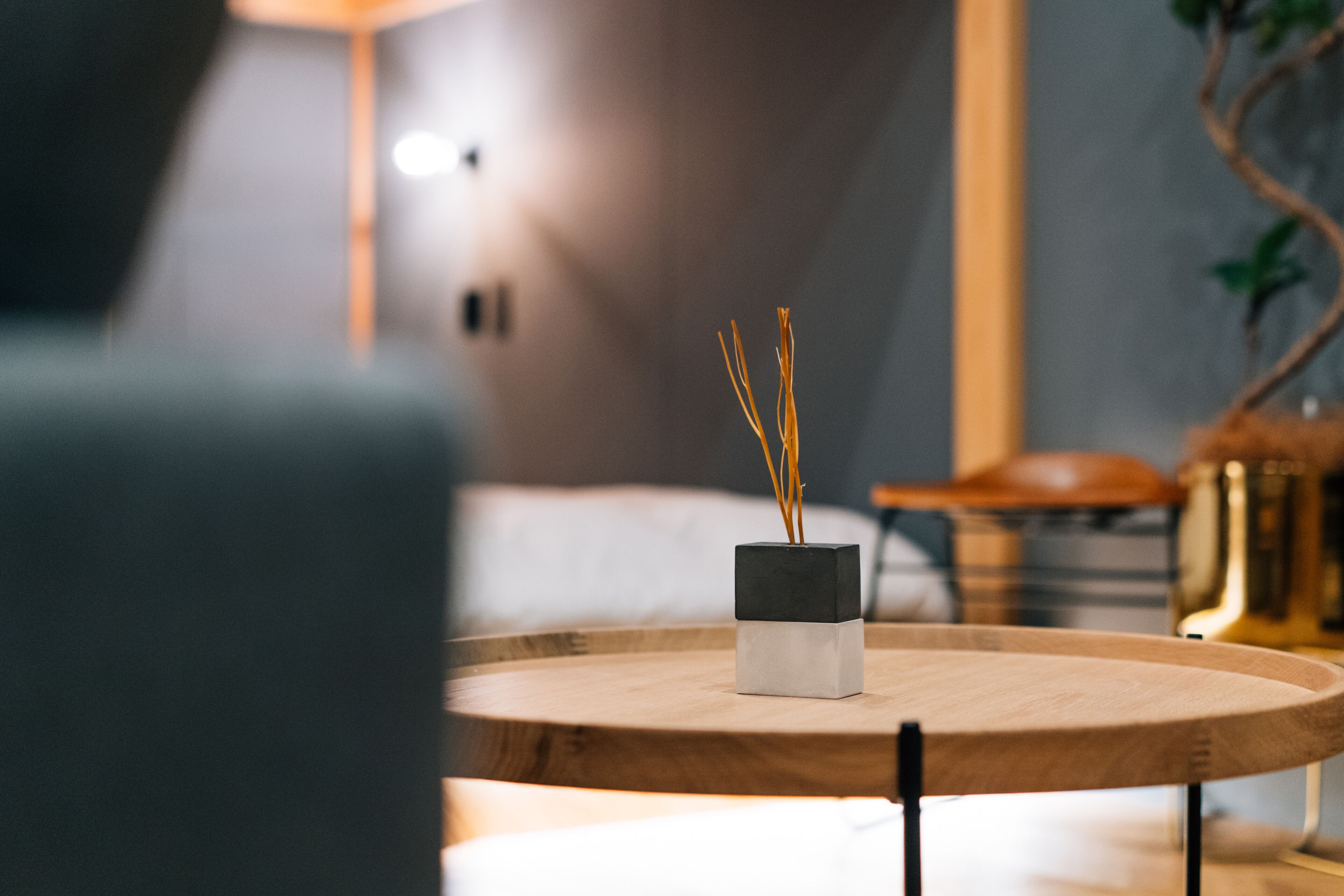 Una habitación moderna con una mesa redonda de madera, un florero blanco y negro con ramas, y una cama al fondo.