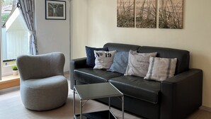 Una sala de estar moderna con un sofá de cuero negro, un sillón gris y una mesa de centro de vidrio. Hay tres obras de arte colgadas en la pared que representan escenas naturales.
