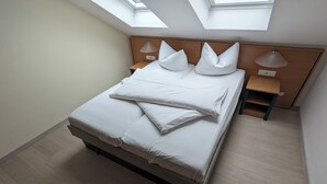 Una cama bien hecha con sábanas y almohadas blancas, cabecera de madera, dos mesitas de noche con lámparas y un tragaluz arriba.
