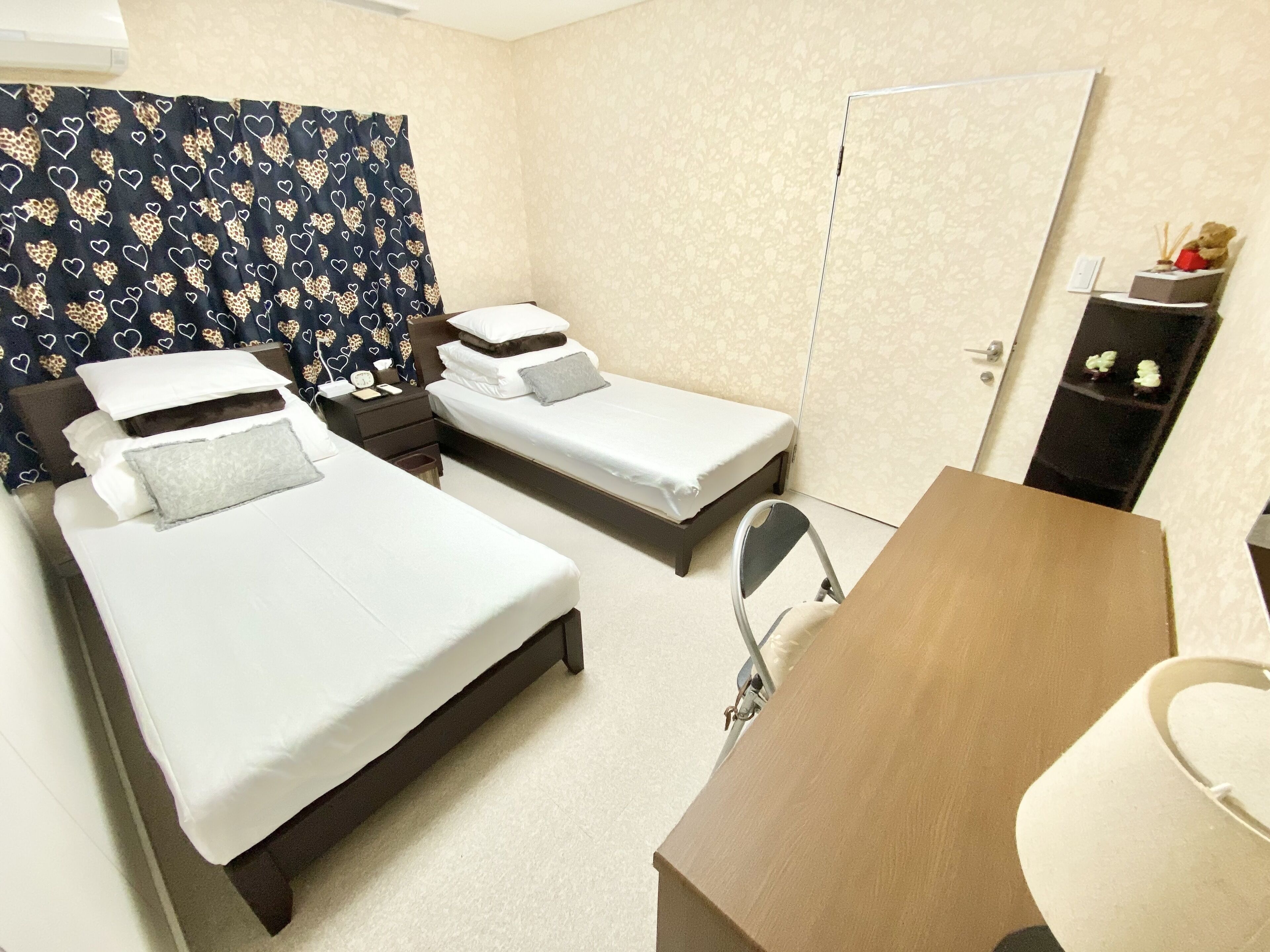 A hotel room with two single beds, a desk, a chair, a wardrobe, and a nightstand with a lamp.