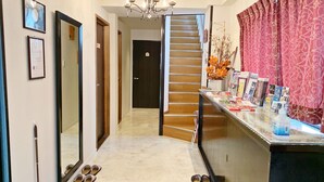 A hallway with a staircase, a mirror, framed certificates, a chandelier, a rug, and a counter with magazines and a bottle. - Guesthouse 017 Reina (Tokushima)
