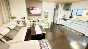 A modern living room with a sofa, a coffee table, and a TV. There is a kitchen area with white cabinets and a microwave. - Guesthouse 017 Reina (Tokushima)
