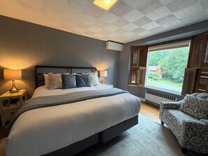 A bedroom with a large bed, bedside lamps, a window with shutters, and a view of greenery outside.