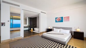 A modern hotel room with a large bed, a view of the sea through a glass door, and a bathroom with a bathtub.