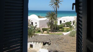 A view through open shutters of a coastal area with white buildings, palm trees, and the sea. - Dar Salem (Mezraia)