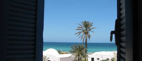 Vista a través de contraventanas abiertas de una zona costera con edificios blancos, palmeras y el mar.