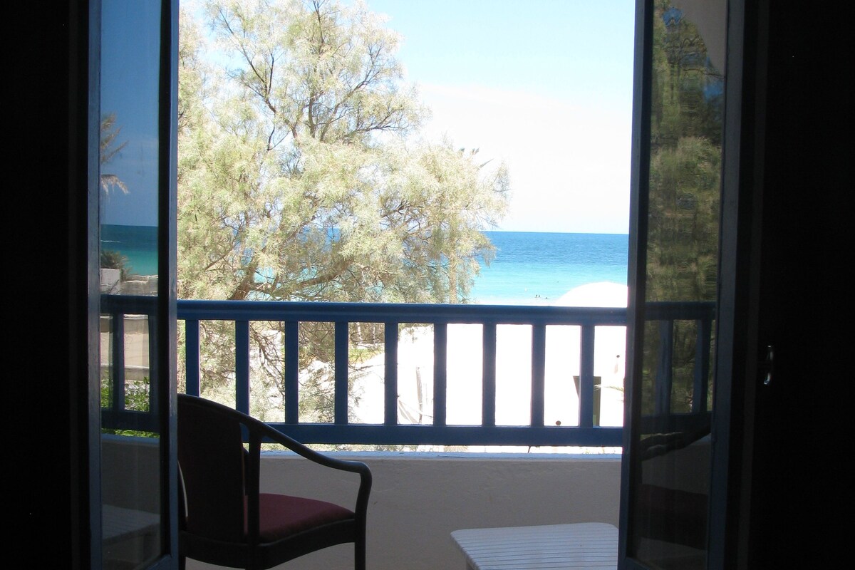 A balcony with a chair and table, overlooking a beach and trees.