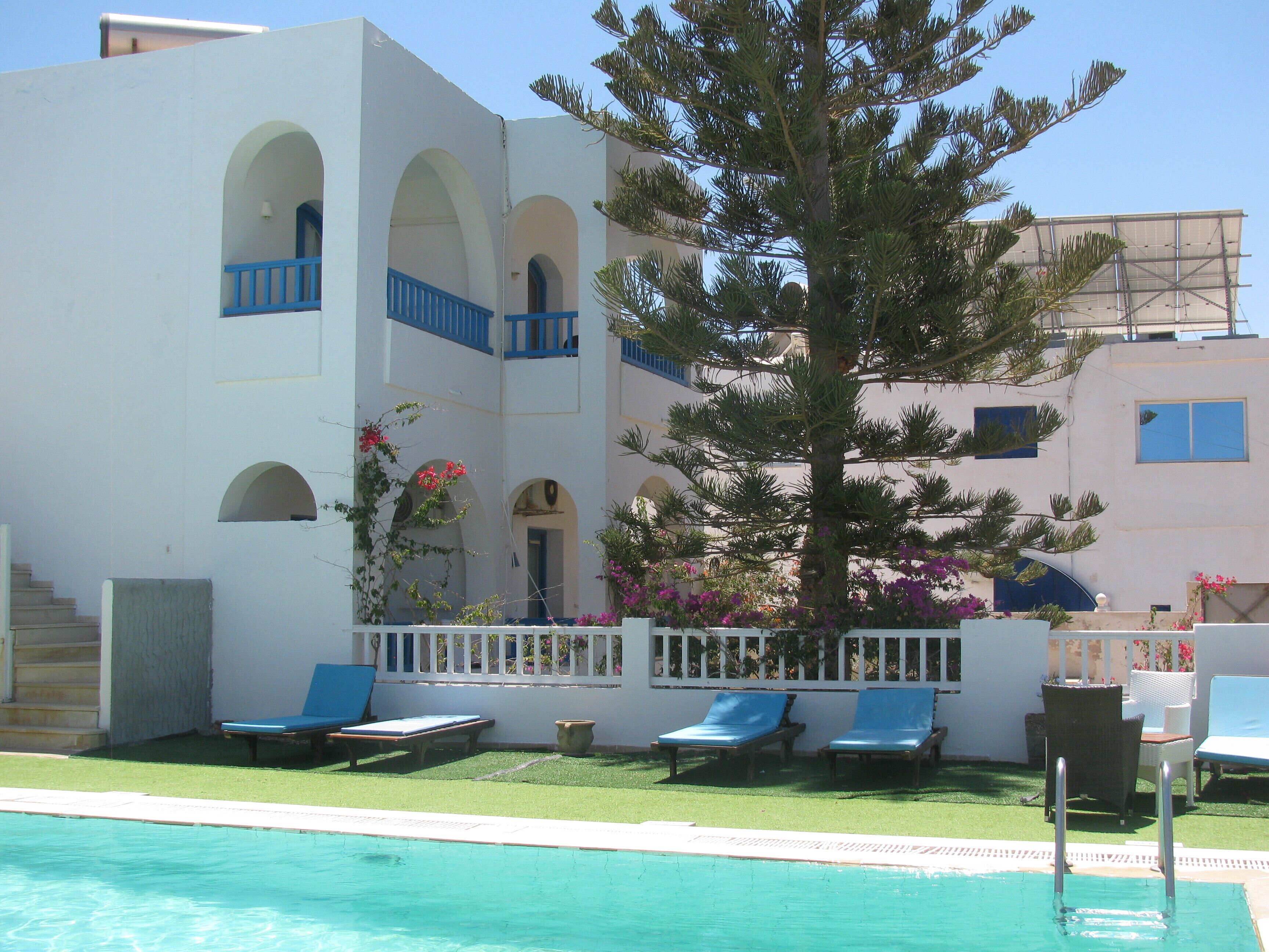 Zona de piscina con un edificio blanco, balcones azules y un árbol grande.