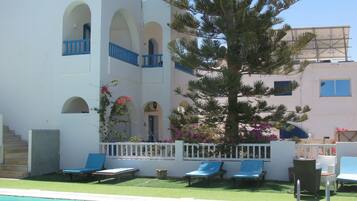 Zona de piscina con un edificio blanco, balcones azules y un árbol grande.