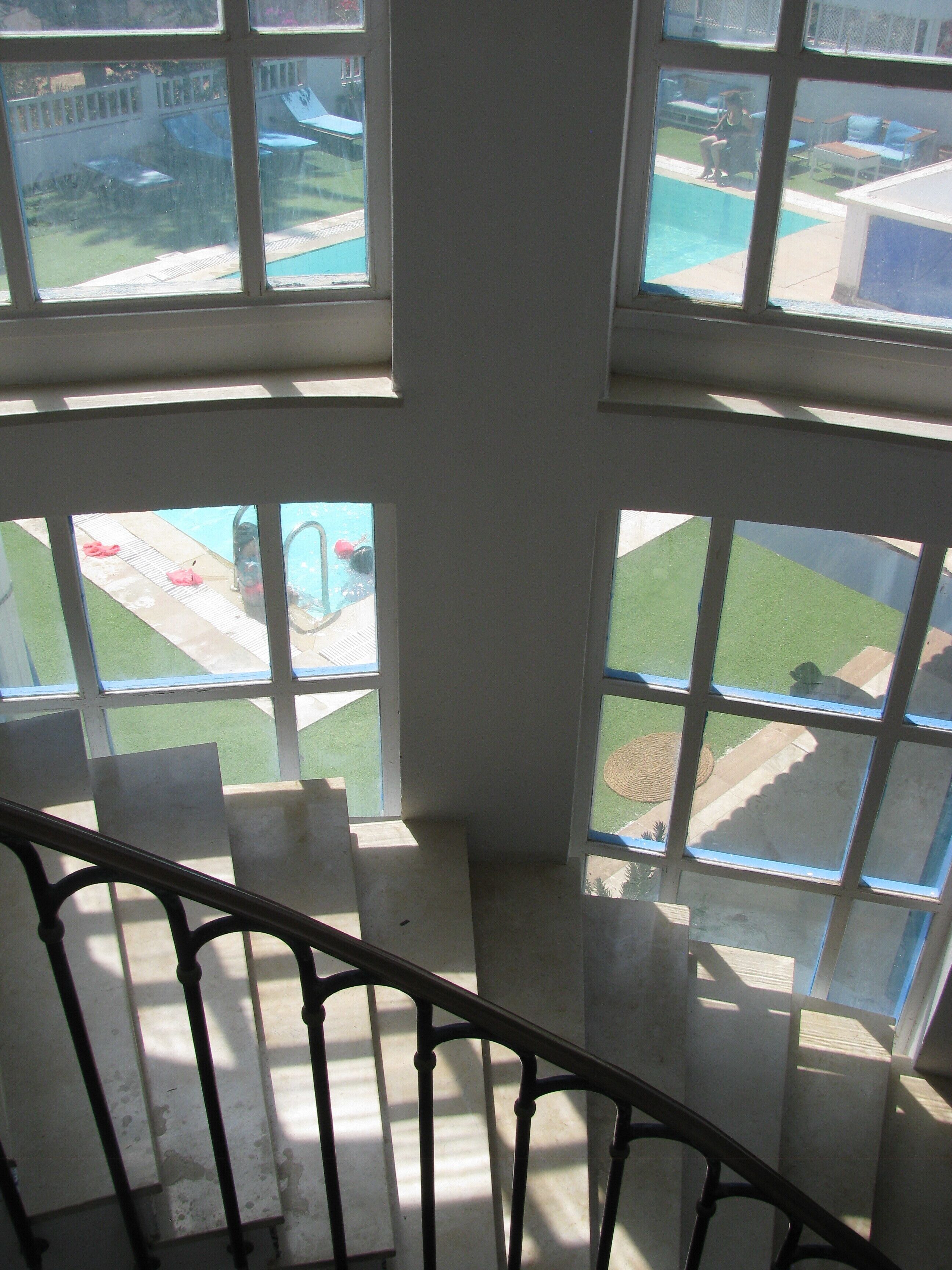 A view from a window with a metal railing, overlooking a swimming pool area with a lawn and a patio with chairs and tables.