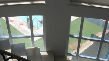 A view from a window with a metal railing, overlooking a swimming pool area with a lawn and a patio with chairs and tables.