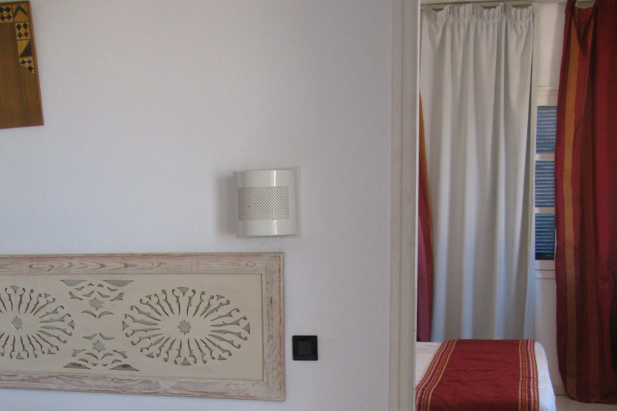 Ein Schlafzimmer mit einem Bett, einem Nachttisch, einer Wandlampe und einem roten Teppich mit Streifen.