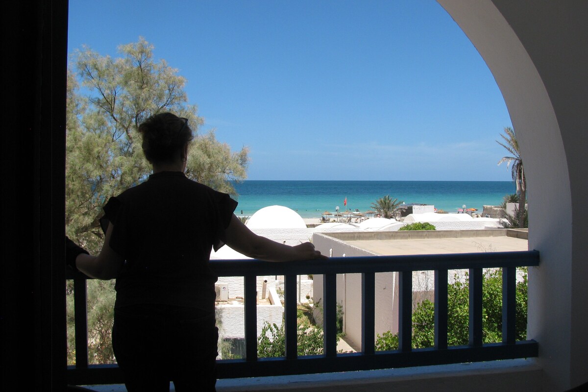 A person standing by a balcony with a view of the sea and a white building in the background.