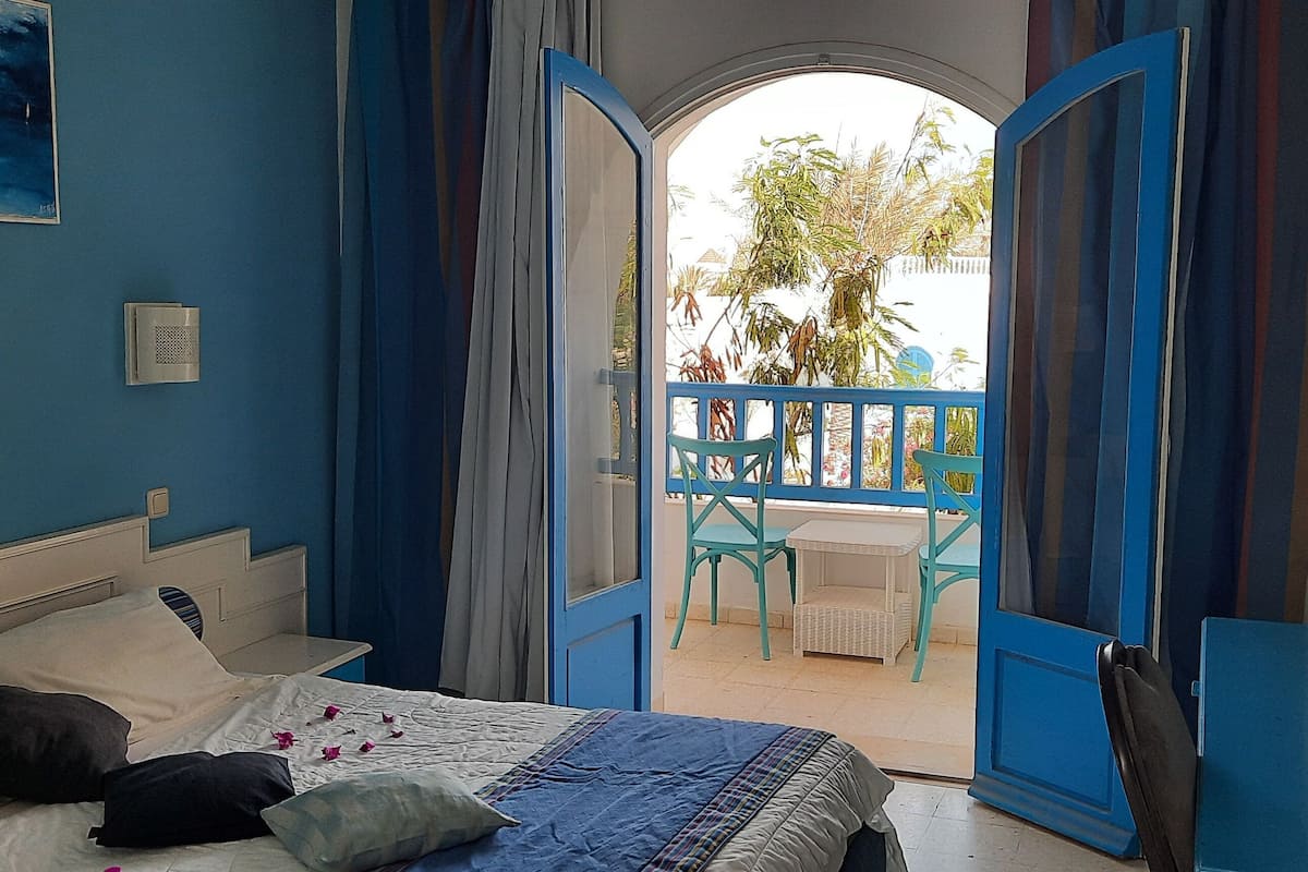 Ein Schlafzimmer mit Bett, Nachttisch, Fernseher und einem Balkon mit Stuhl und Pflanzen.