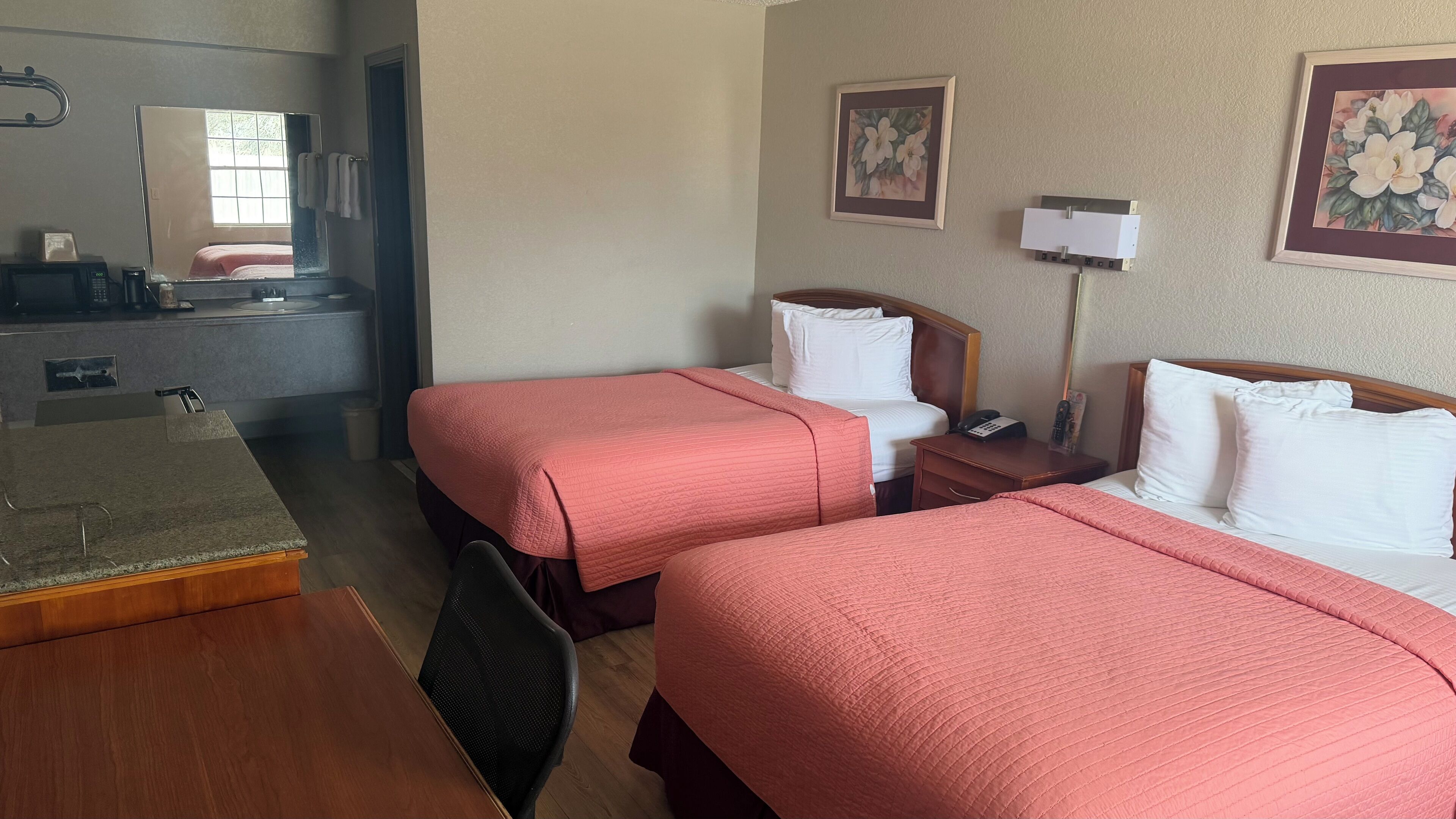 A hotel room with two beds, a desk, a mirror, a small table, and a wall-mounted lamp.