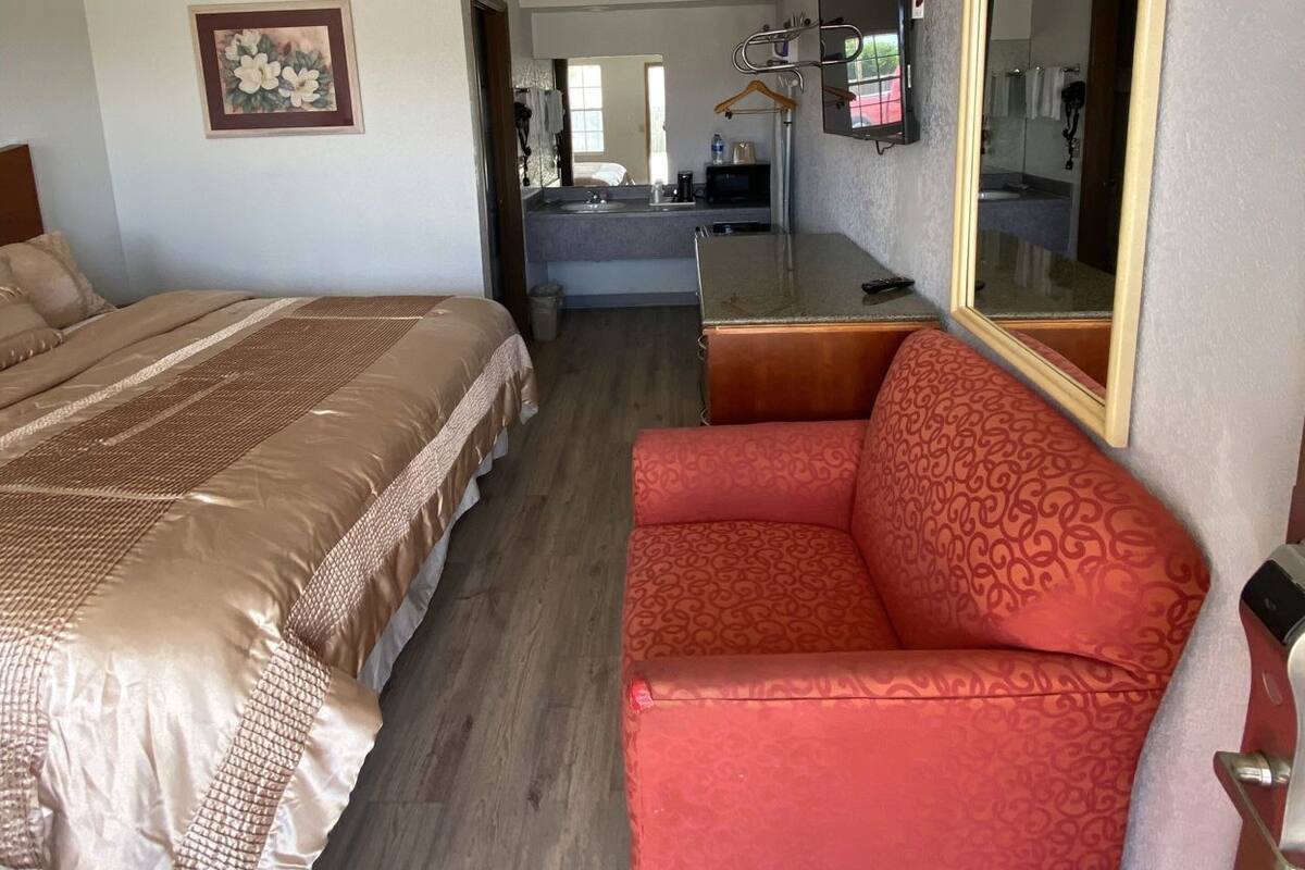 A hotel room with a bed, a red armchair, a desk with a mirror, and a painting on the wall.