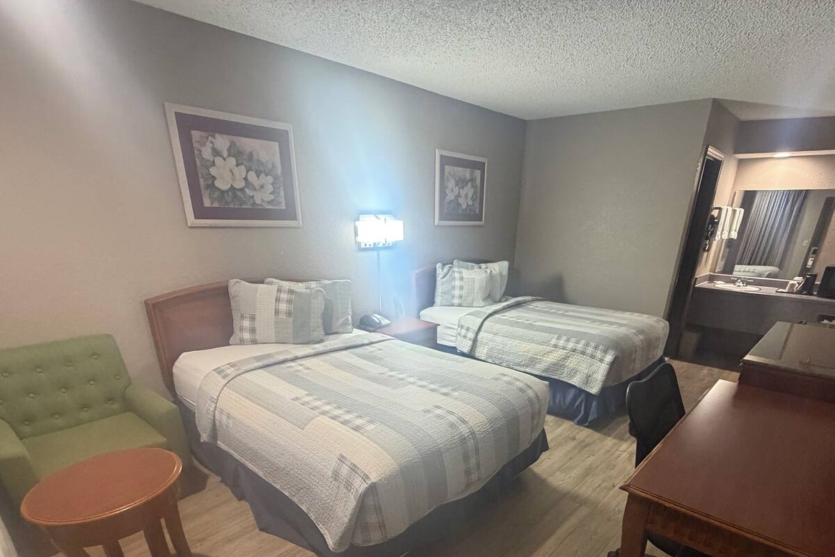 A hotel room with two beds, a green armchair, a wooden table, and a desk with a chair.
