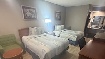 A hotel room with two beds, a green armchair, a wooden table, and a desk with a chair.