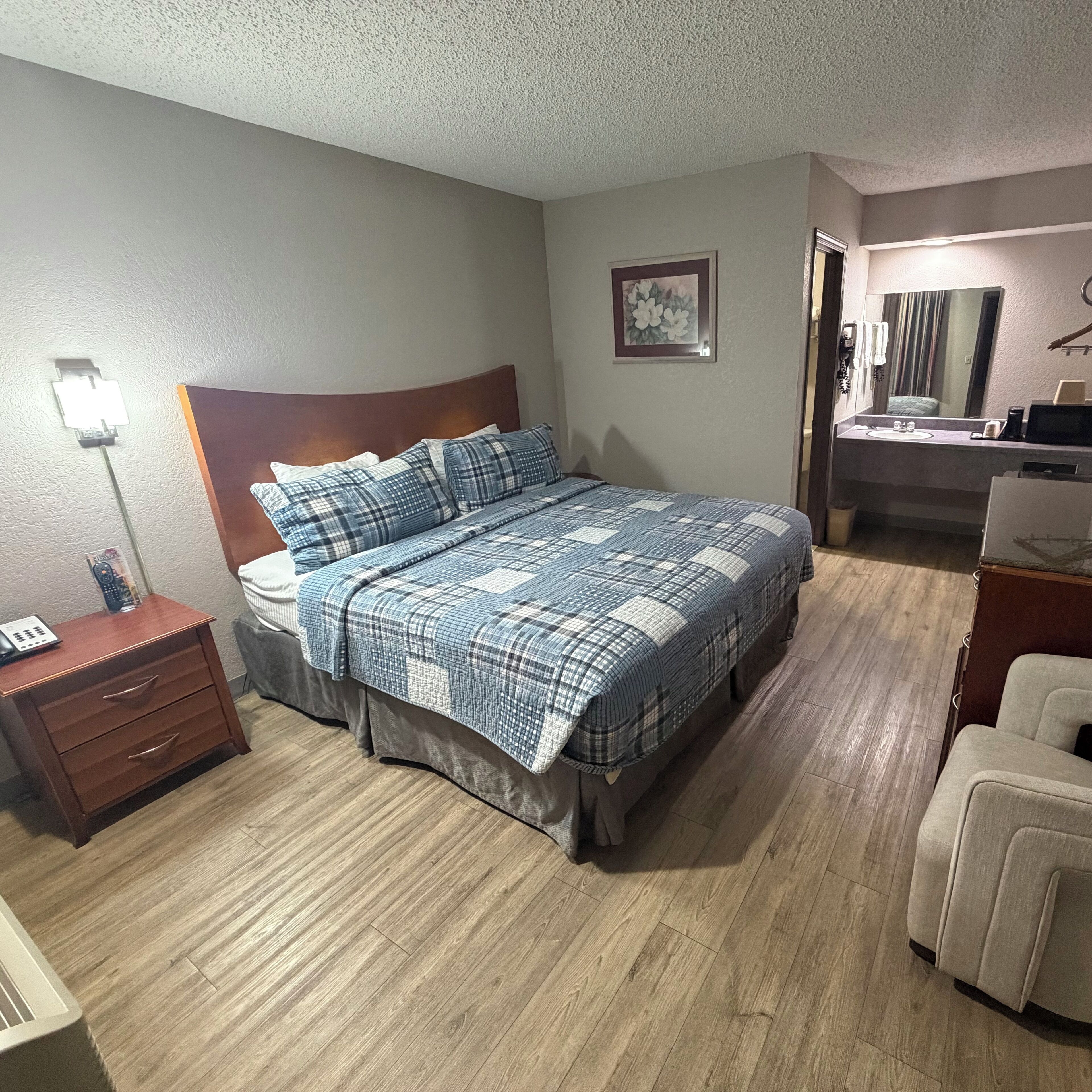A bedroom with a bed, a nightstand, a dresser, a chair, a bathroom with a sink and mirror, and a painting on the wall.