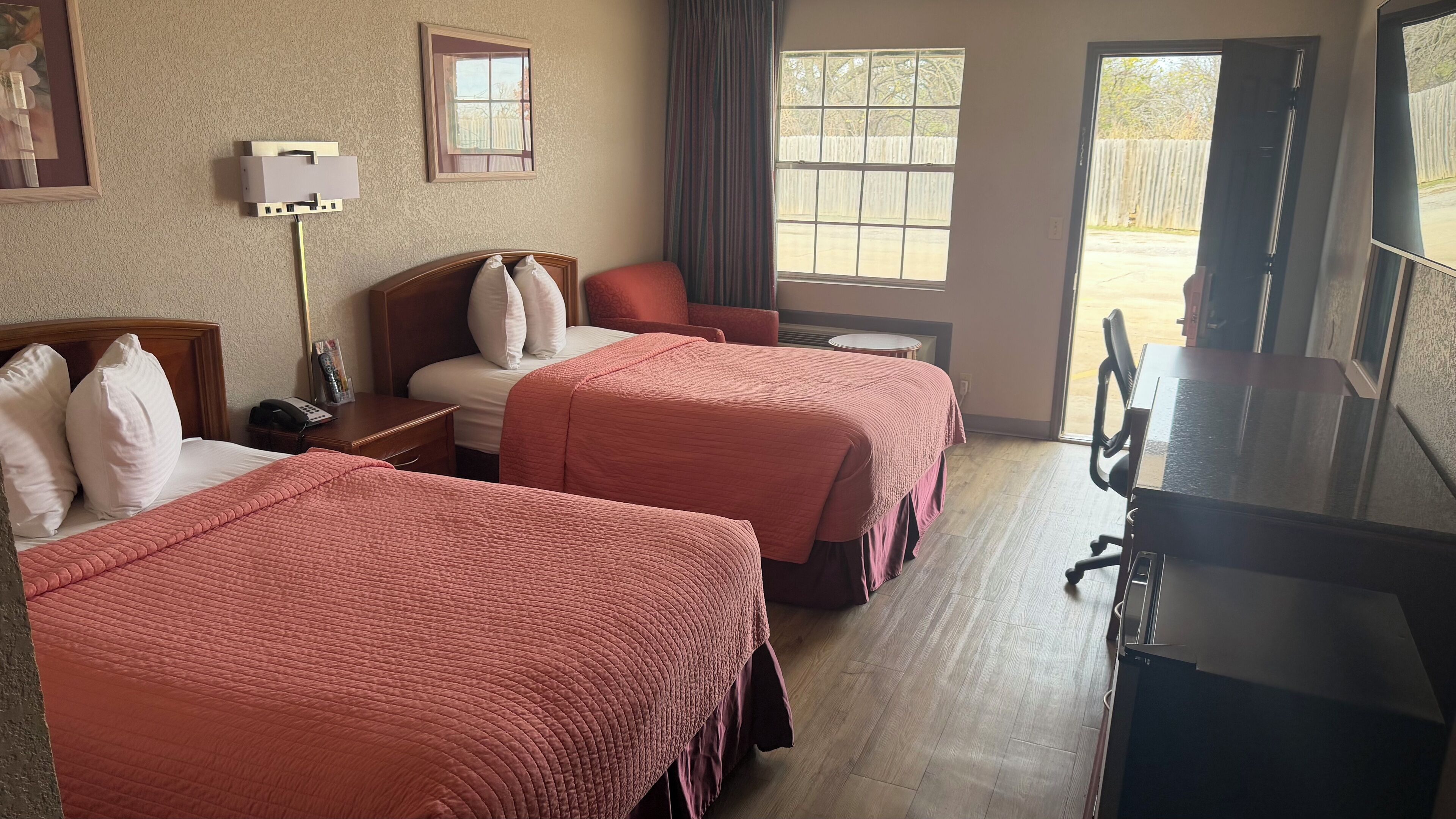 A hotel room with two beds, a desk, a chair, a TV, and a window with a view of a fence.