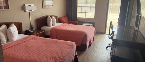 A hotel room with two beds, a desk, a chair, a TV, and a window with a view of a fence.