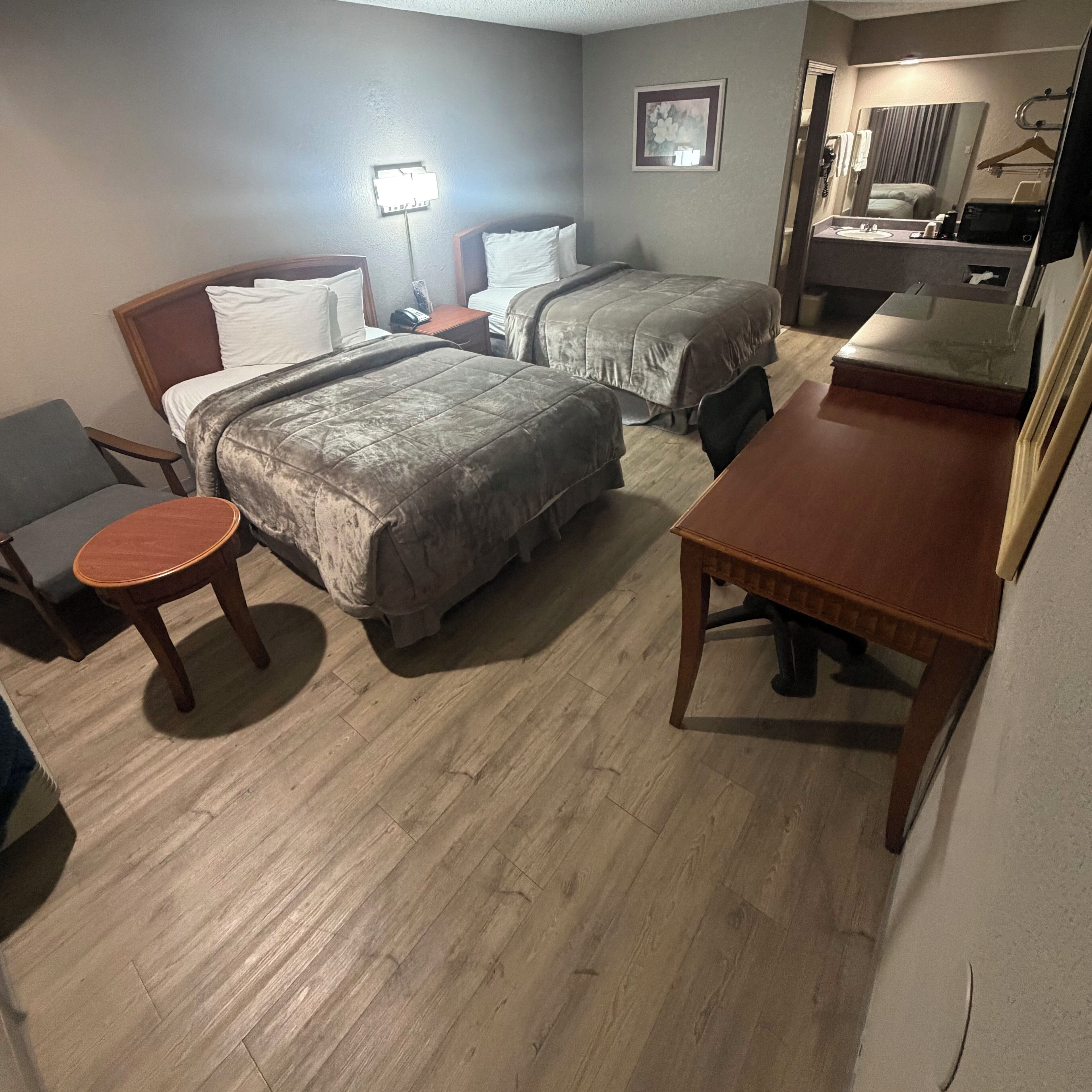 Habitación de hotel con dos camas, una mesita redonda pequeña, un escritorio de madera, un espejo y un baño con lavamanos y espejo.
